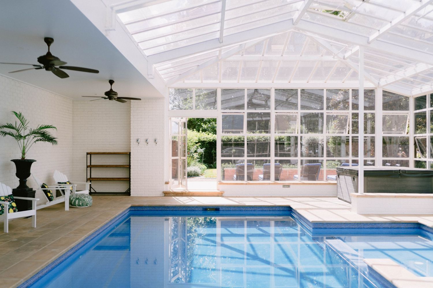 Indoor swimming pool surrounded by glass walls and white ceiling with ceiling fans and potted plants beside the pool.