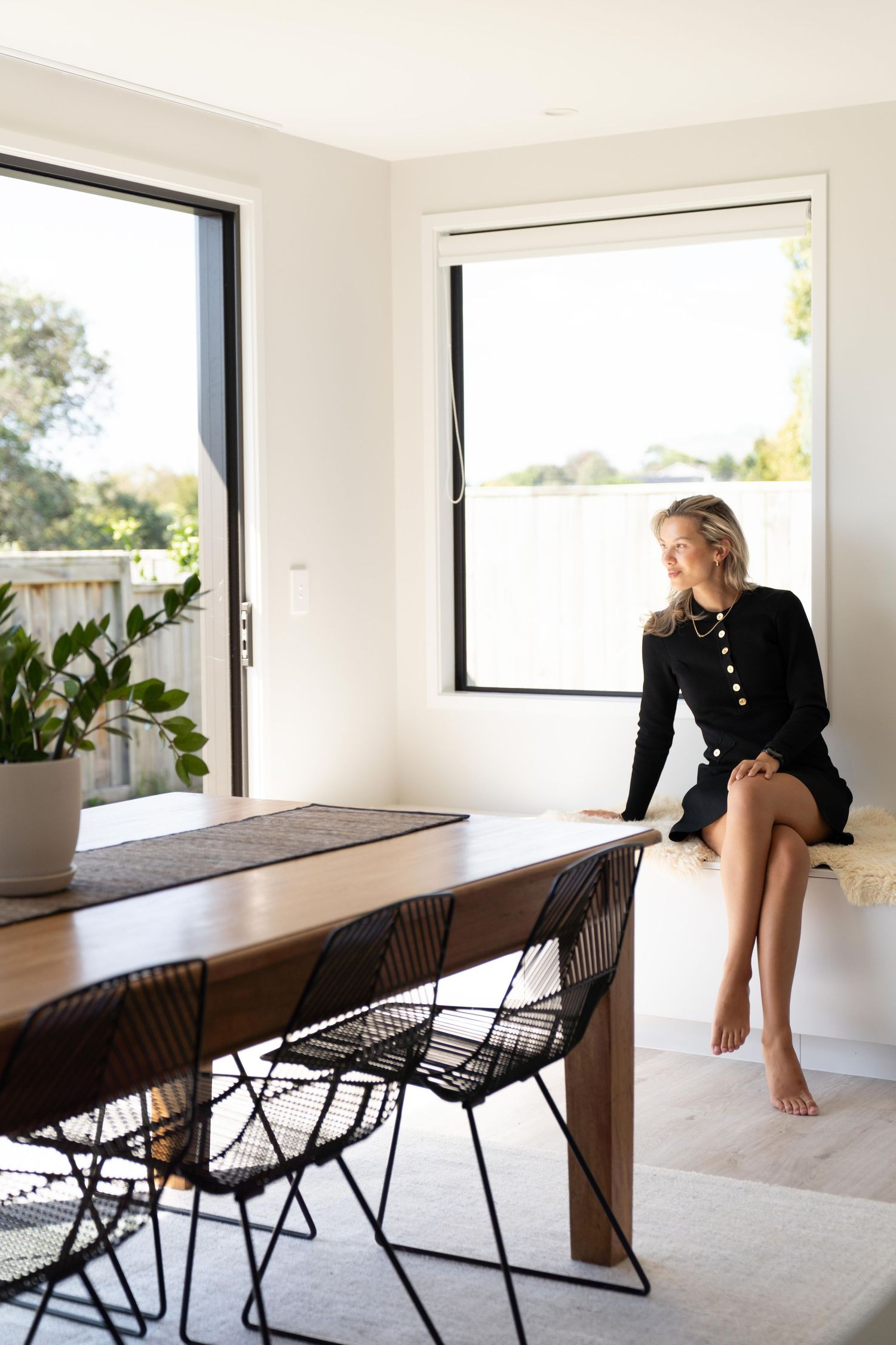 Modern dining room with wooden table, black wire chairs, and figure sitting on window ledge in black dress.