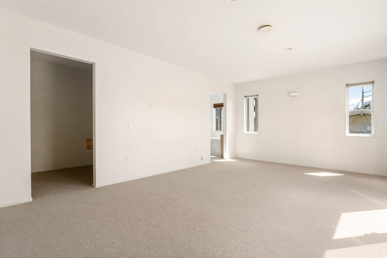 Large empty room with grey carpet flooring, white walls and natural light from windows.