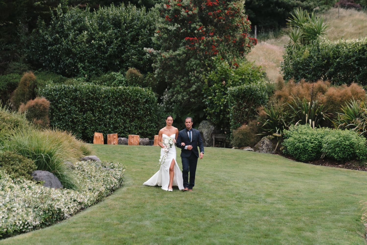 A couple walks through a lush garden setting surrounded by flowering shrubs and green landscaping during an outdoor celebration.
