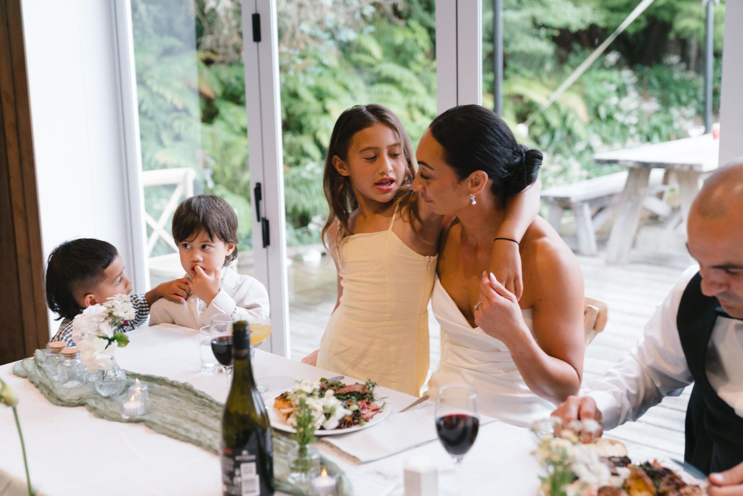 Wedding reception guests enjoying a meal inside a bright, windowed venue.