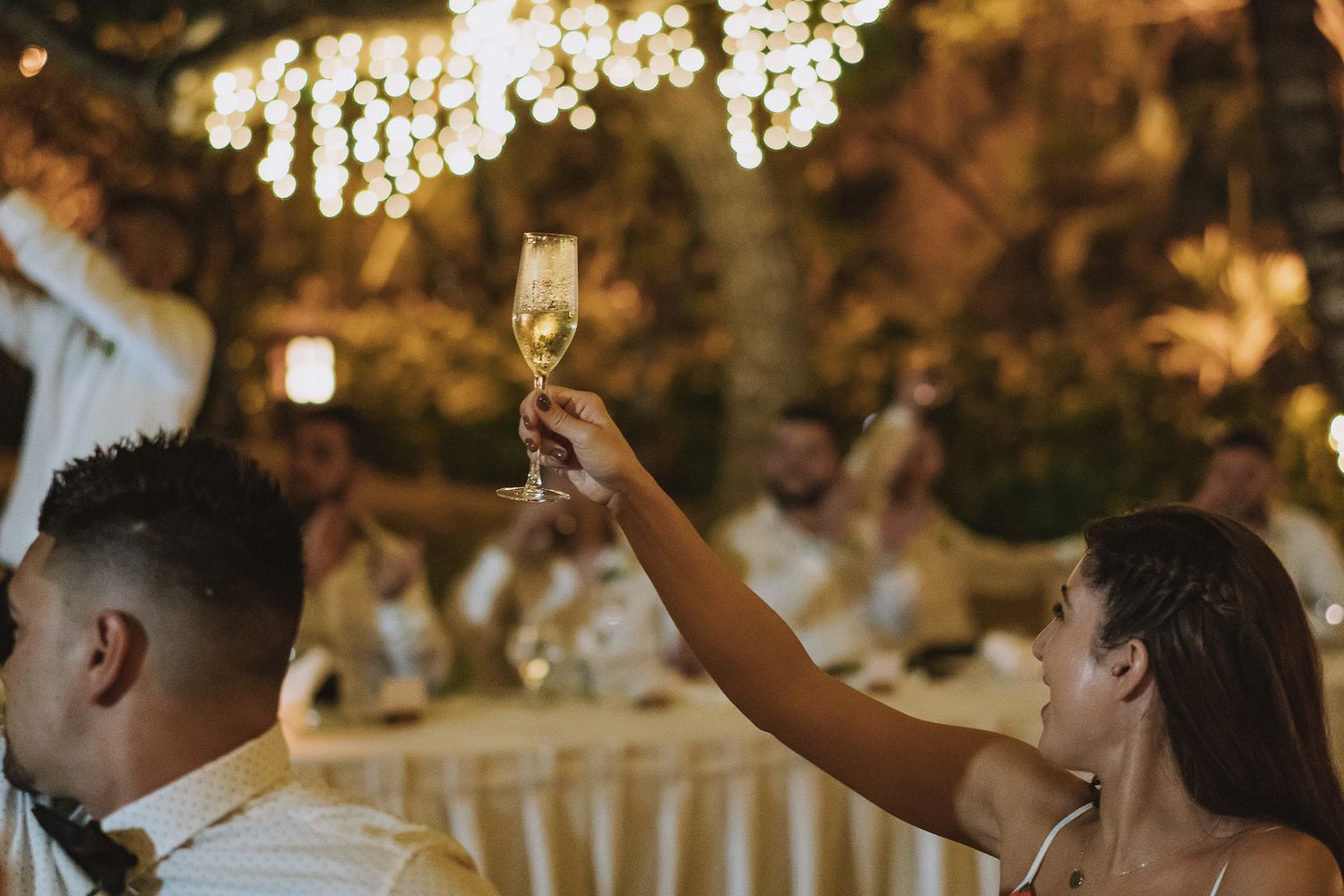 People raise champagne glasses in celebration under glowing string lights at an elegant outdoor wedding reception.