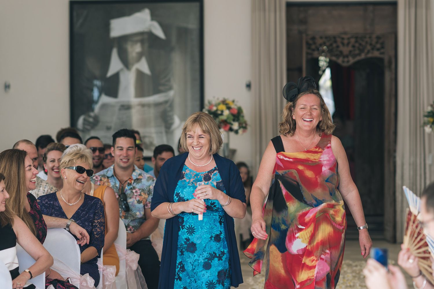 Wedding guests in colorful attire walk down an aisle with joyful expressions.
