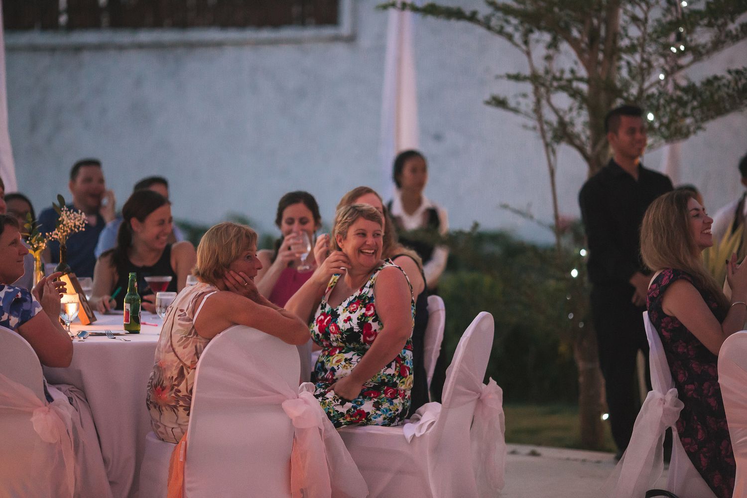 Wedding guests seated at tables under white tents during an evening outdoor reception.