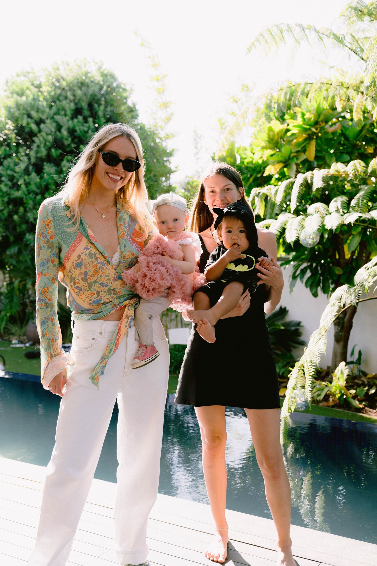 Several people gather outdoors by a pool on a sunny day sharing a playful moment together in light summer attire.