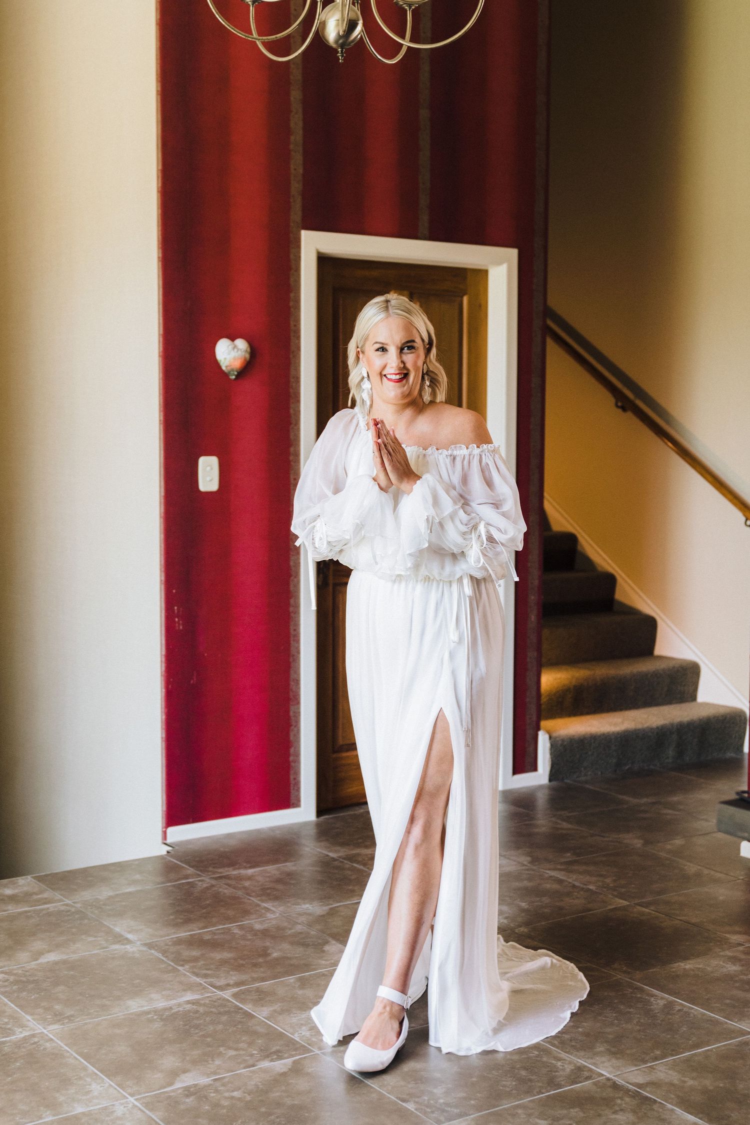 Elegant off-shoulder white wedding dress with flowing fabric and high slit shown in foyer with red accent wall.