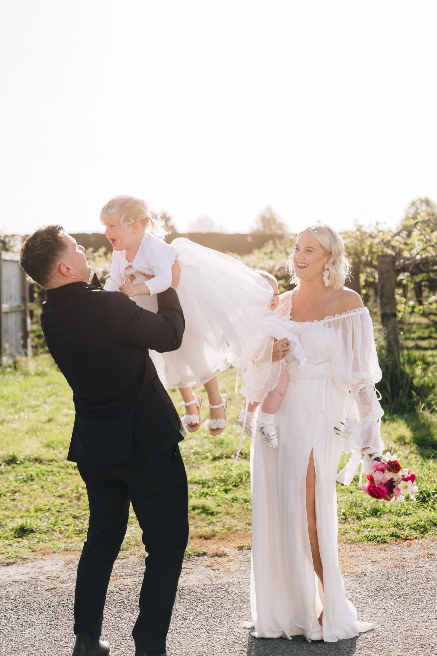 Happy family moment at a wedding as parents lift their small child while standing outdoors in formal attire.