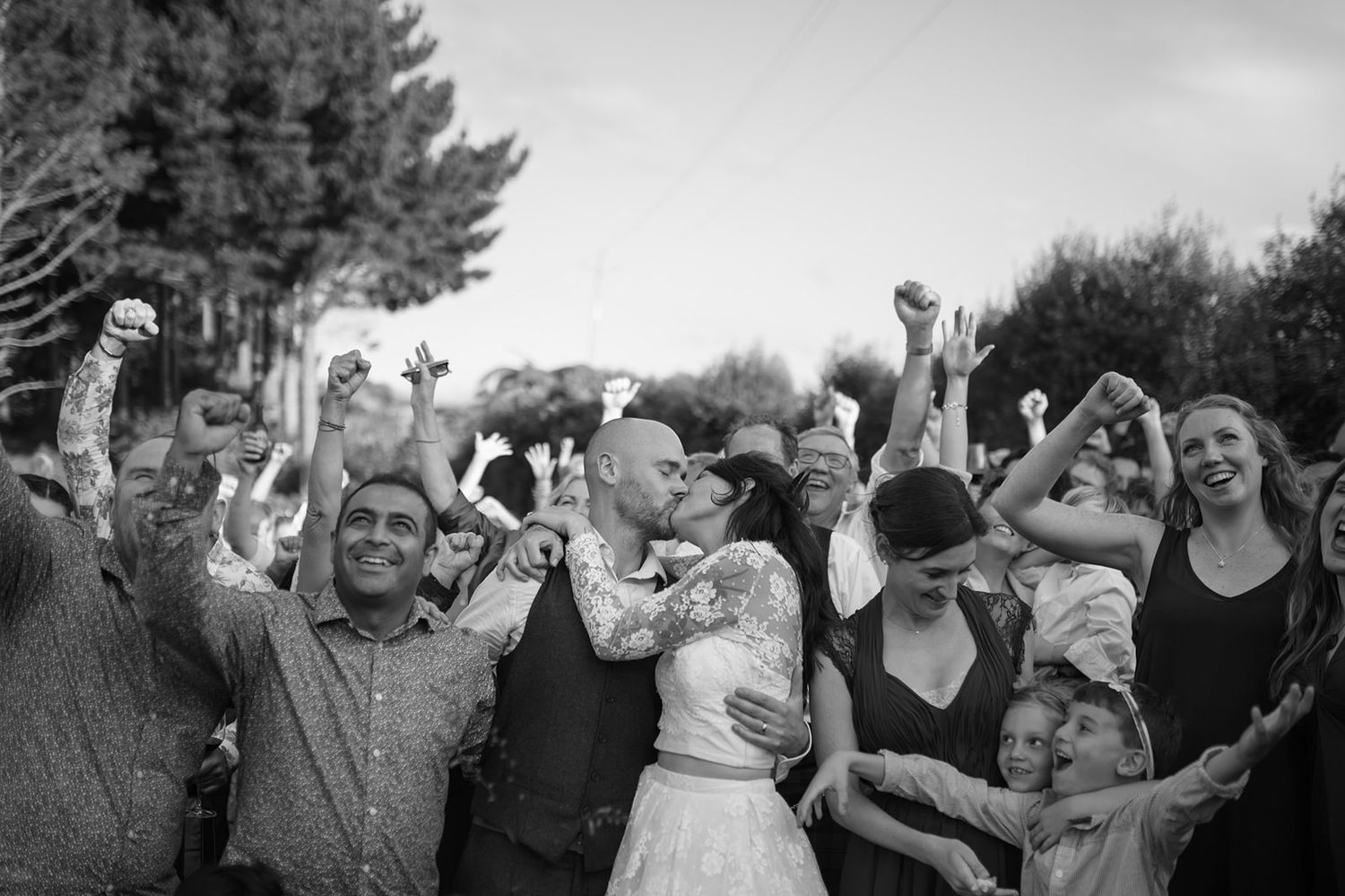 A joyful crowd celebrates at an outdoor wedding reception with arms raised and cheers in black and white.