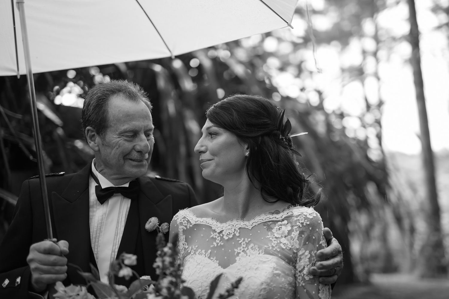 Black and white photo of a couple sharing an umbrella outdoors, with the bride wearing lace and groom in a bowtie.