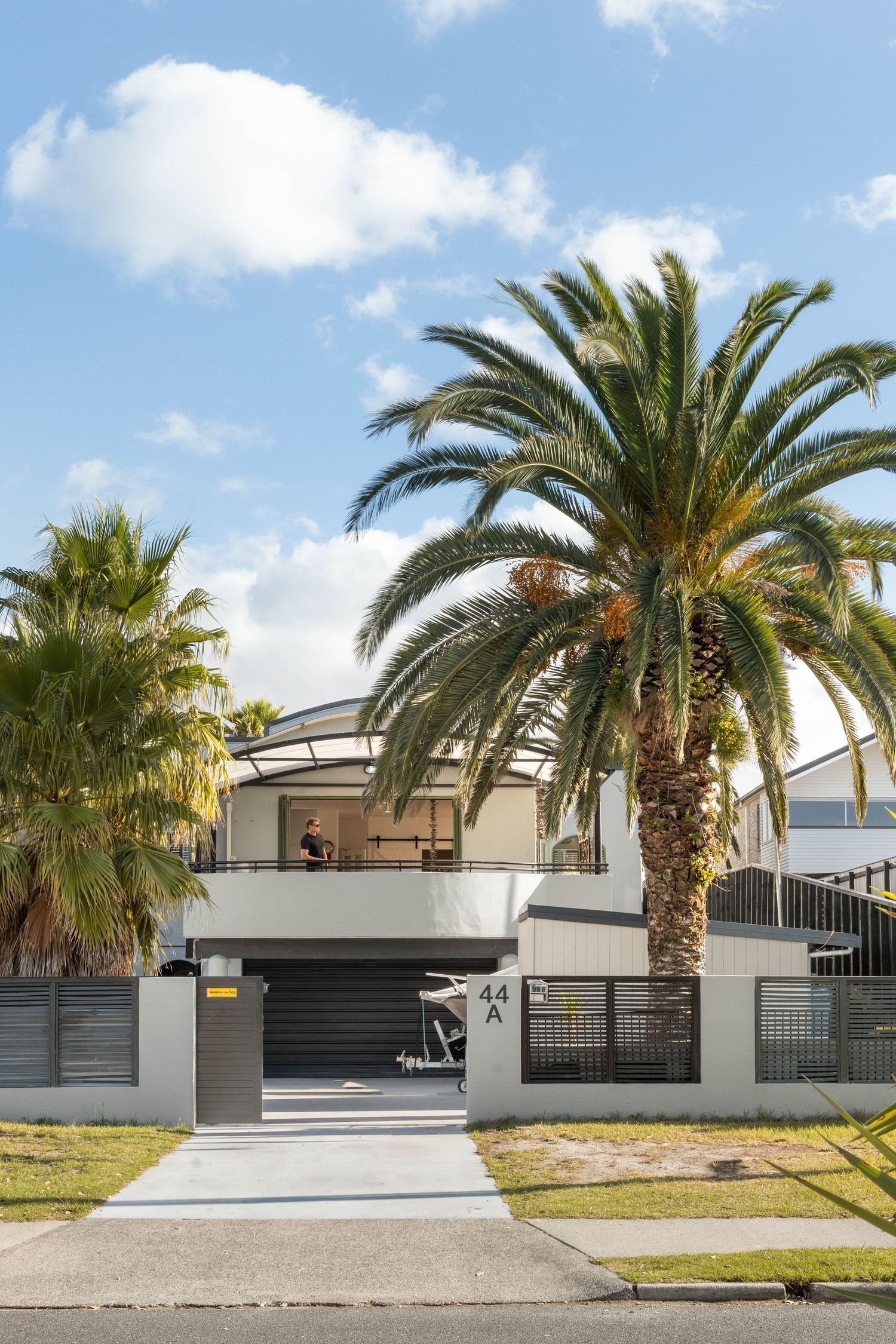 Modern white house exterior with palm trees and black gates along a paved driveway.