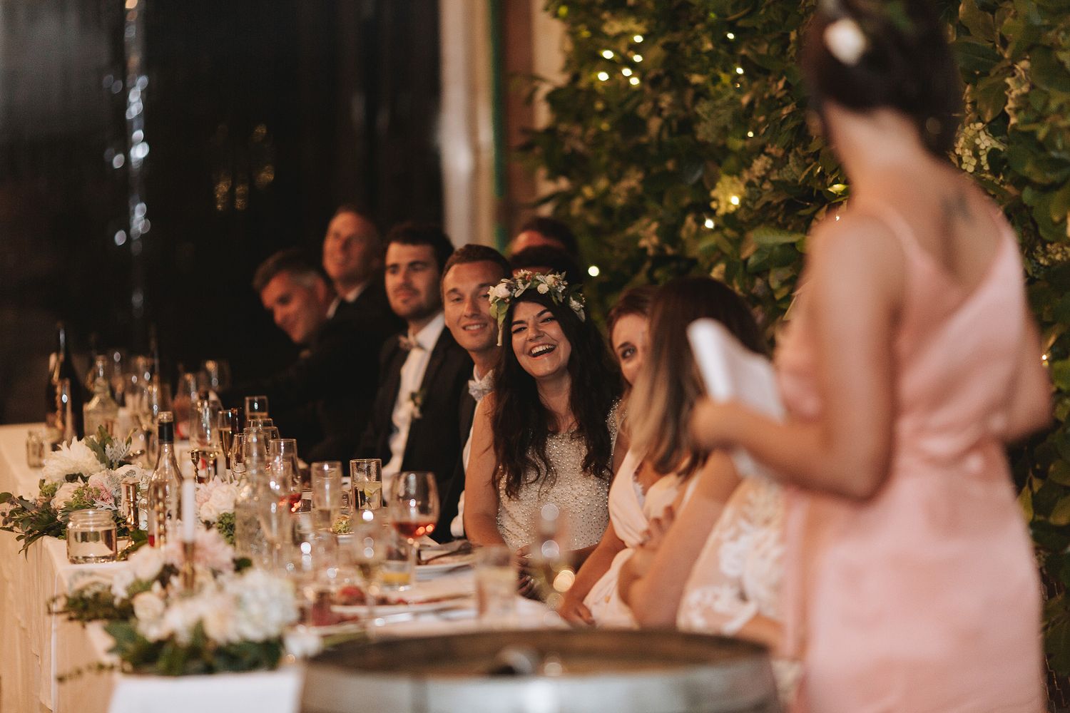 Wedding reception guests smile and laugh together at an elegant candlelit dinner table with pink floral centerpieces.