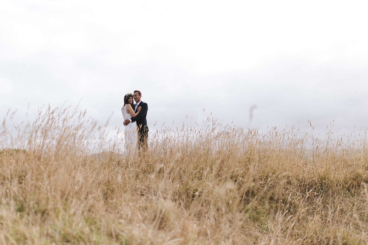 A couple embraces in a field of tall golden grass on a misty day, creating a romantic silhouette against the sky.
