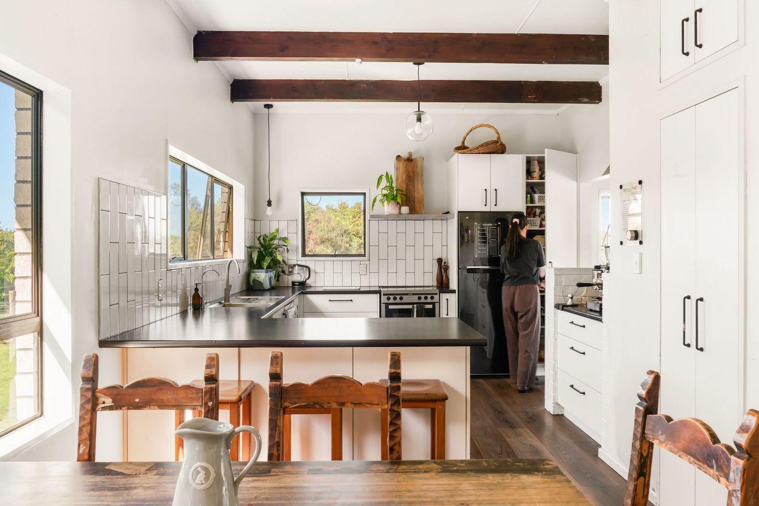 Bright modern kitchen with exposed wooden beams, white cabinets, and rustic wooden dining chairs by a breakfast bar.