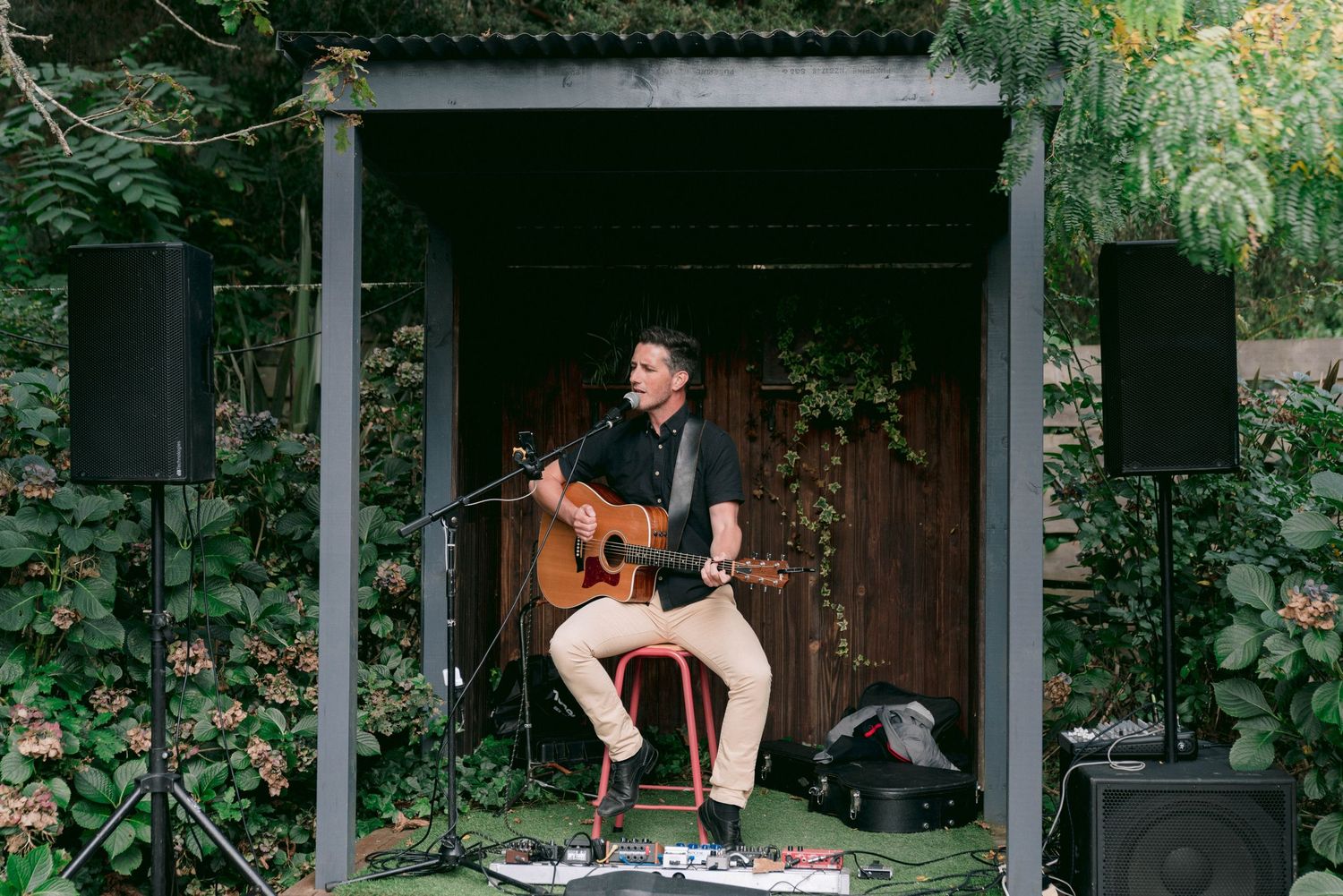 Guitarist performs acoustic set outdoors in cozy garden gazebo setting surrounded by lush greenery and vines.