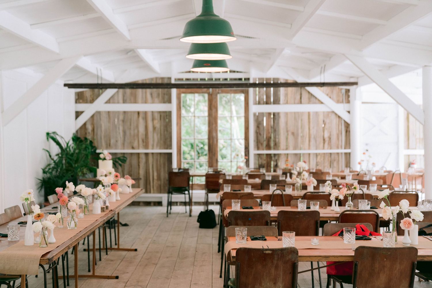 Rustic wedding reception venue with wooden tables, green chairs and pink floral centerpieces under vaulted white ceiling.