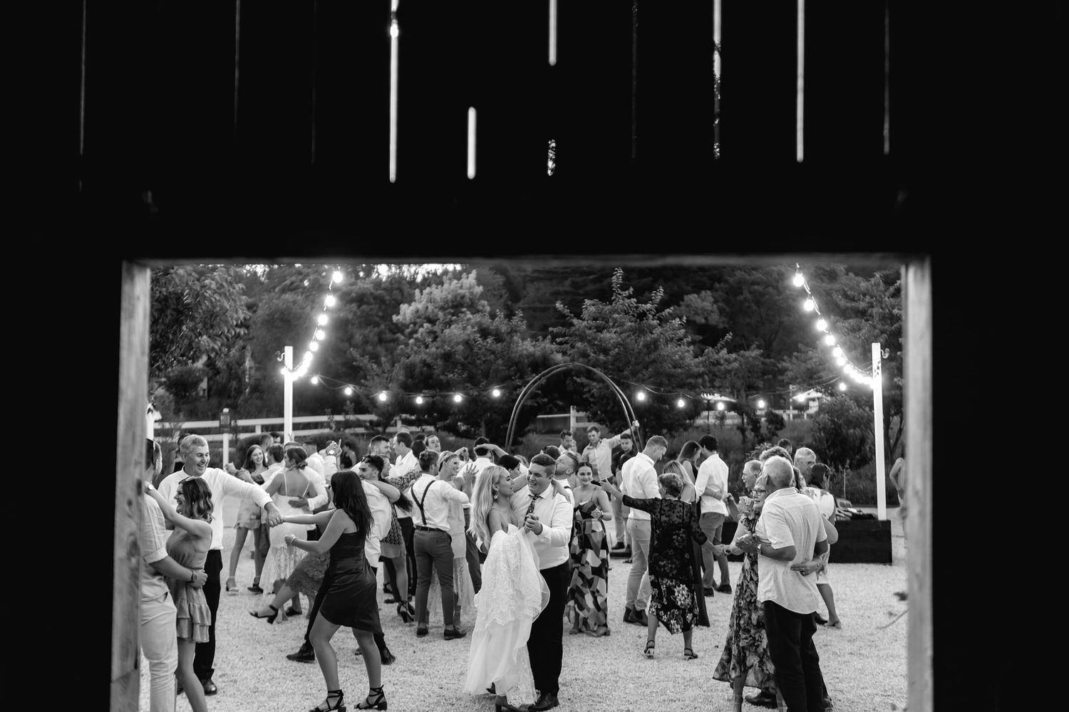 A black and white photograph shows people dancing outdoors under strung lights at an evening social gathering.