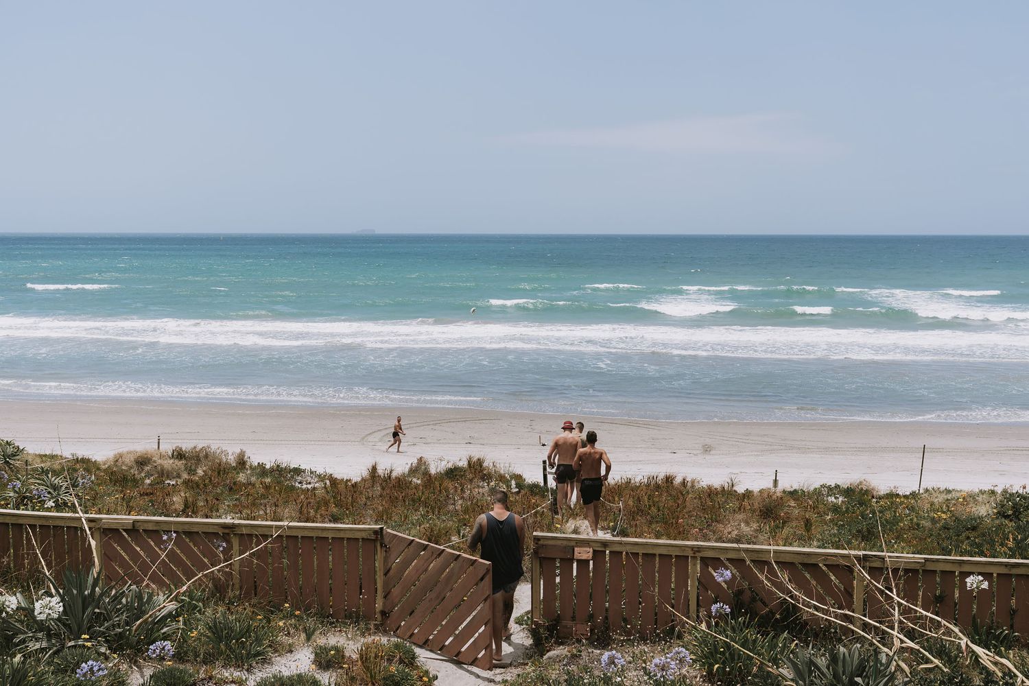 Two people descend wooden stairs to a pristine beach with turquoise waves crashing on white sand.