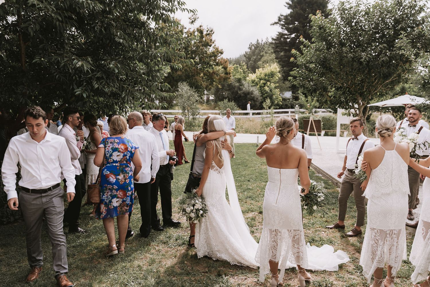 Outdoor summer wedding celebration with guests gathered on a grassy lawn under trees on a sunny afternoon.