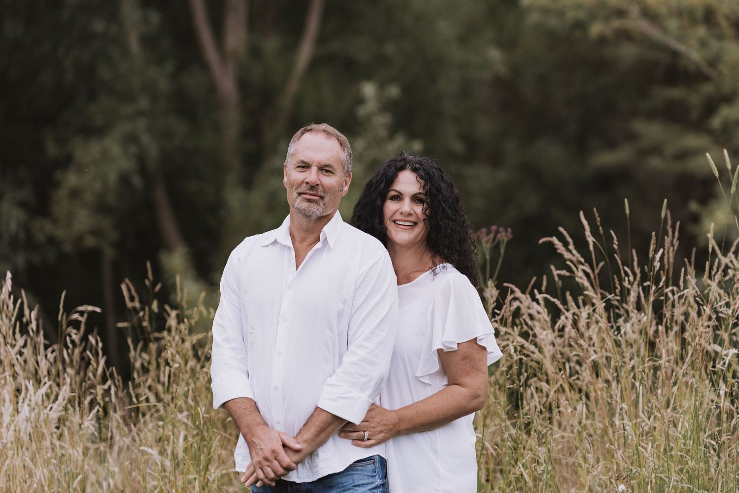 A couple embraces while sitting in a sunlit meadow with tall grass and trees in the background.