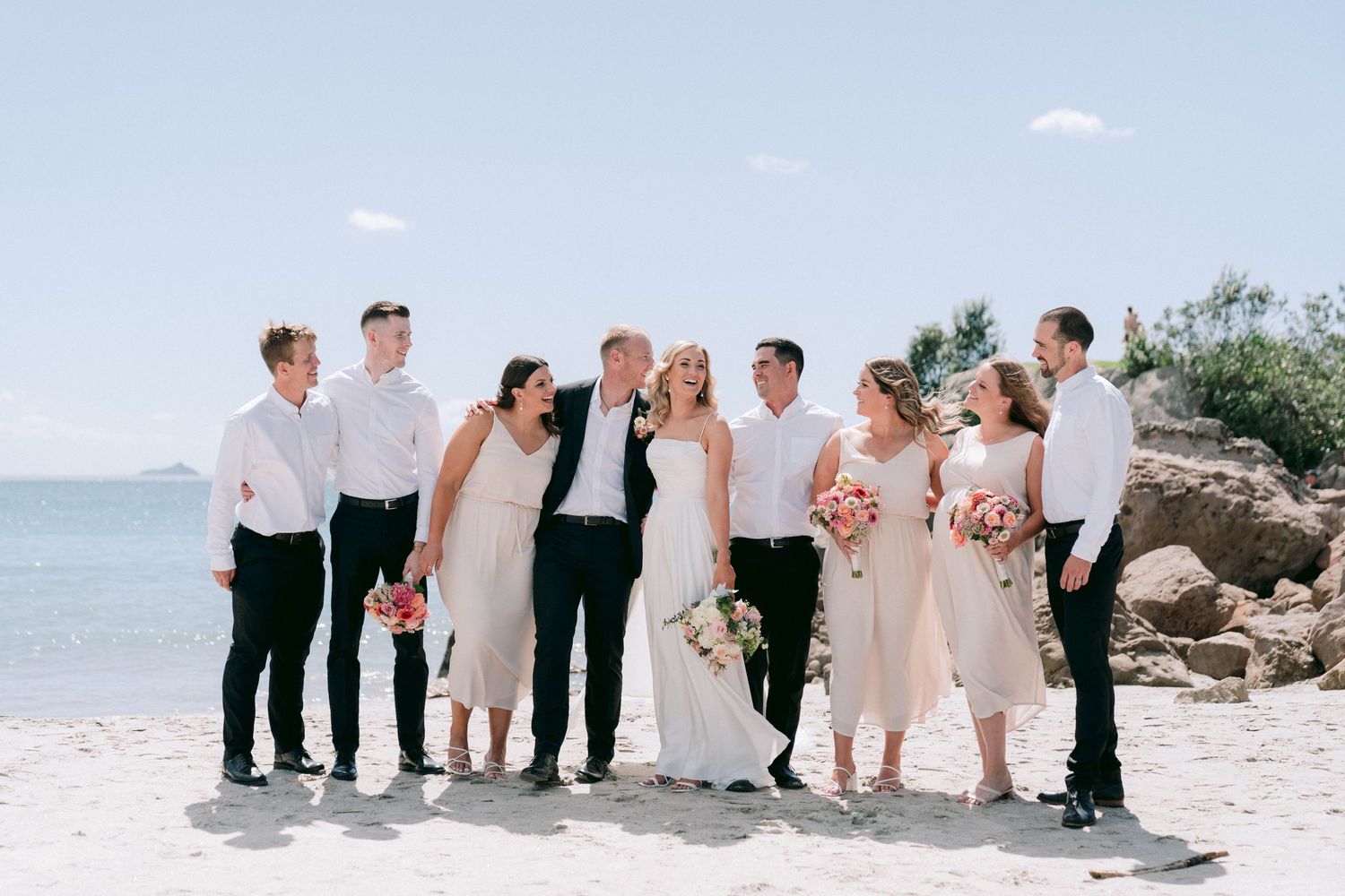 Wedding party wearing light colors poses together on a sunny beach with ocean views in the background.