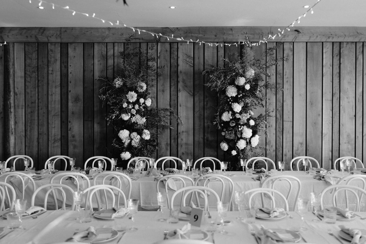 Black and white wedding reception setup with bentwood chairs and floral arrangements against a wooden wall.