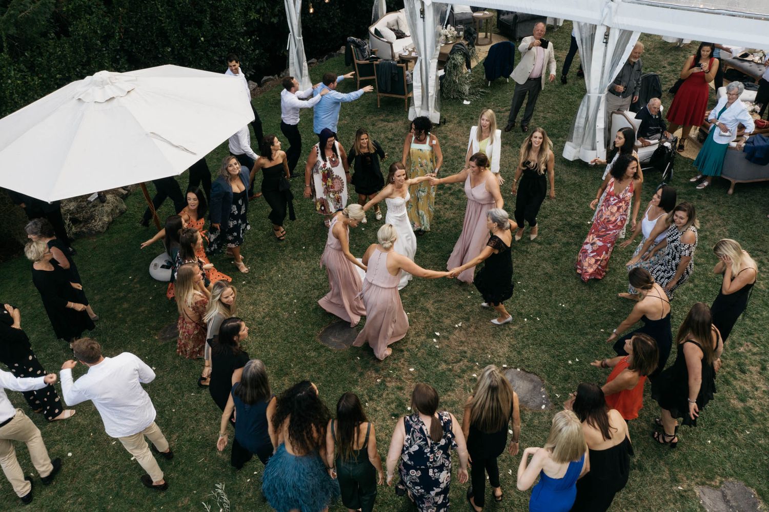 A group of people dancing in a circle on a grass lawn under a white tent during an outdoor celebration.