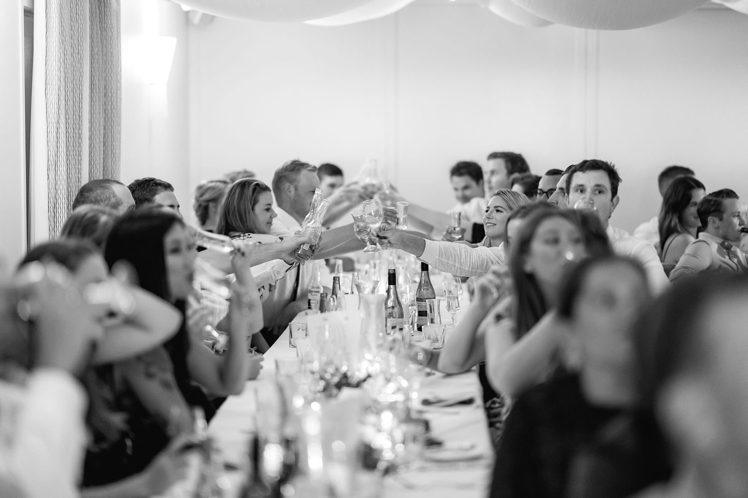 Wedding reception guests raise their glasses in a toast at a long banquet table during an elegant celebration.