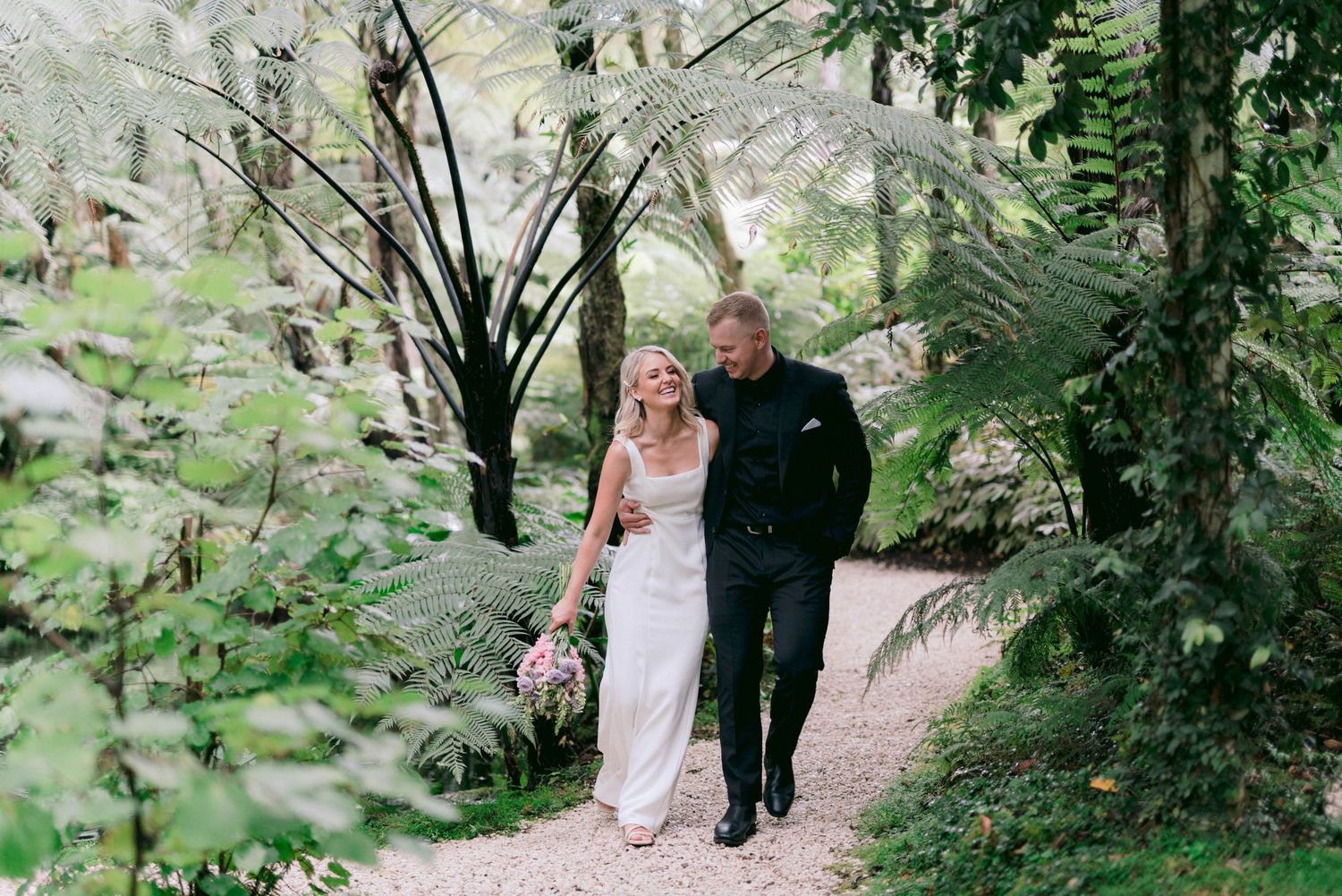 A couple strolls along a garden path surrounded by lush greenery and ferns.