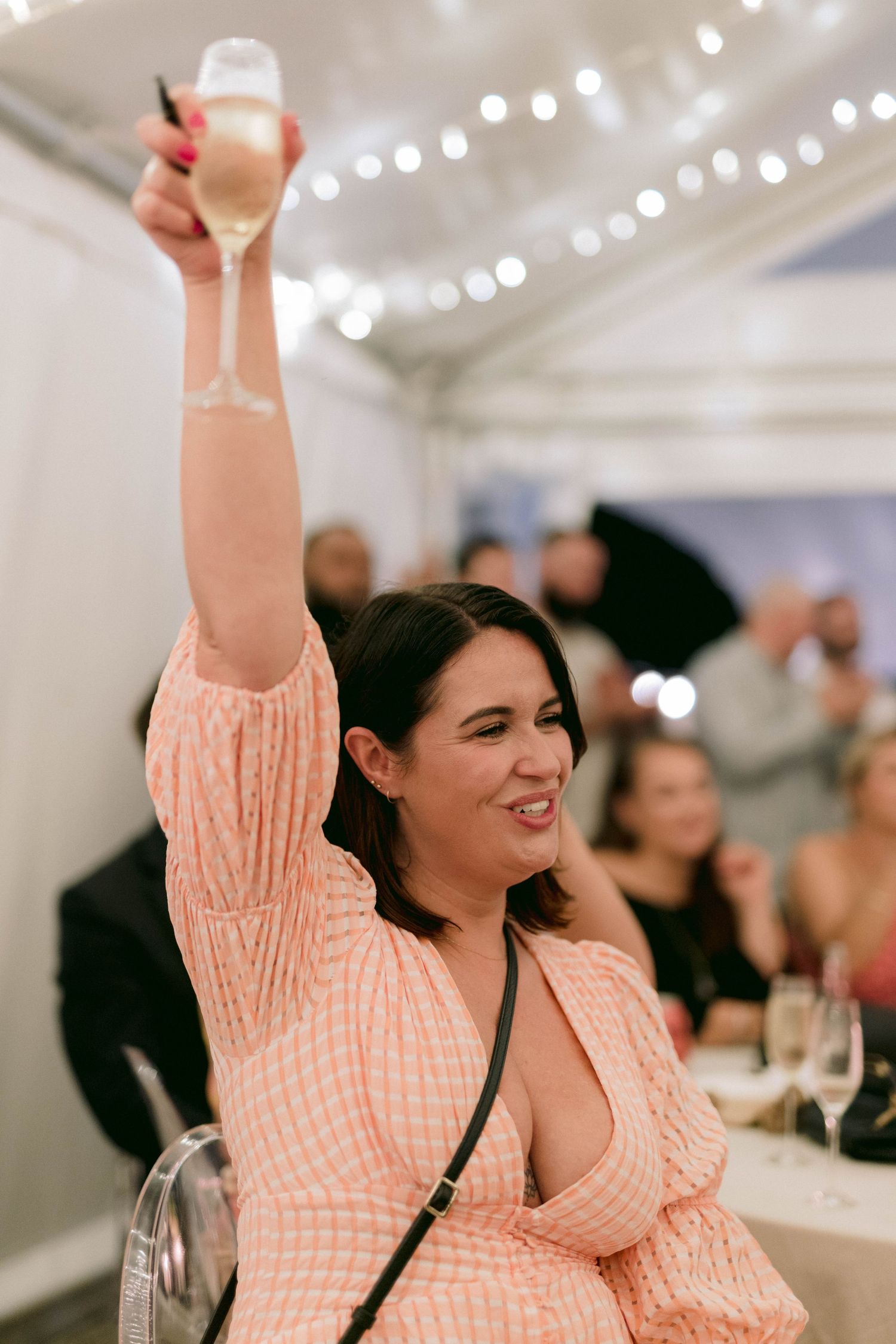 A guest in a peach dress raises their champagne glass in celebration at a wedding reception.