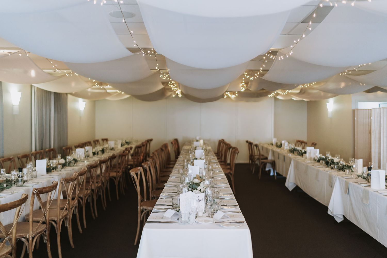 Elegant banquet hall setup with long rows of white-clothed tables, white draped ceiling, and soft mood lighting.