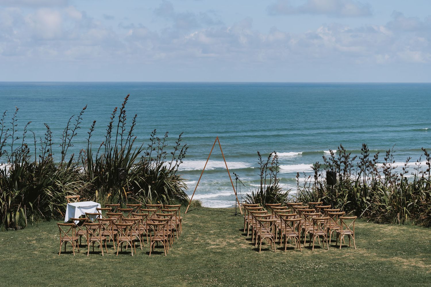 Wooden chairs set up for a beach wedding ceremony with ocean views and natural coastal vegetation in the background.
