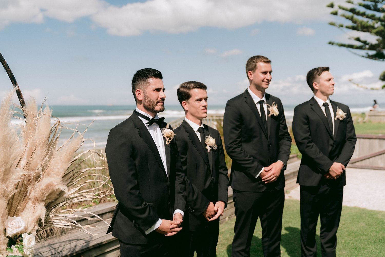 Groomsmen in black tuxedos and suits stand together at a scenic oceanside wedding ceremony location.