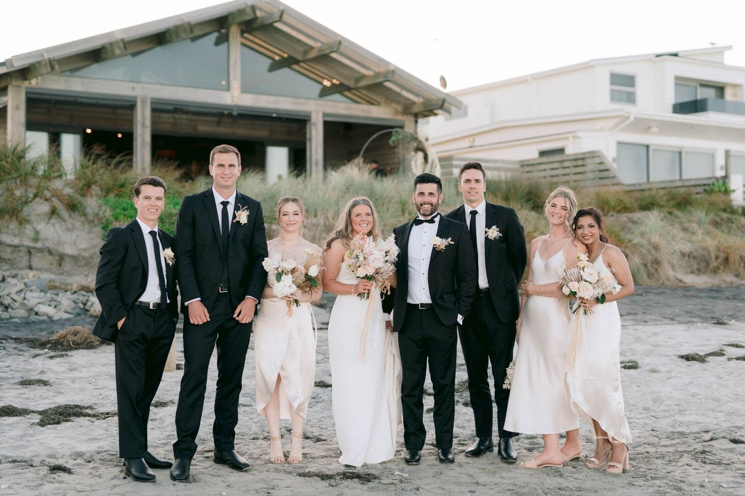 Wedding party in formal attire poses on beach with modern building in background.