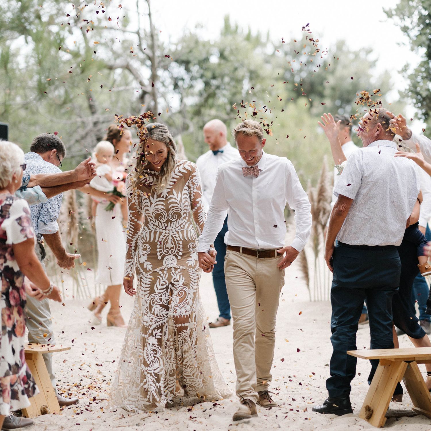 Wedding guests celebrate by throwing confetti as the newlyweds walk through an outdoor garden path lined with trees.