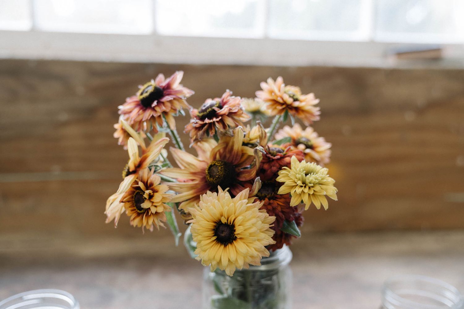 A rustic arrangement of dried sunflowers and autumn blooms in glass jars lined up on a wooden surface.