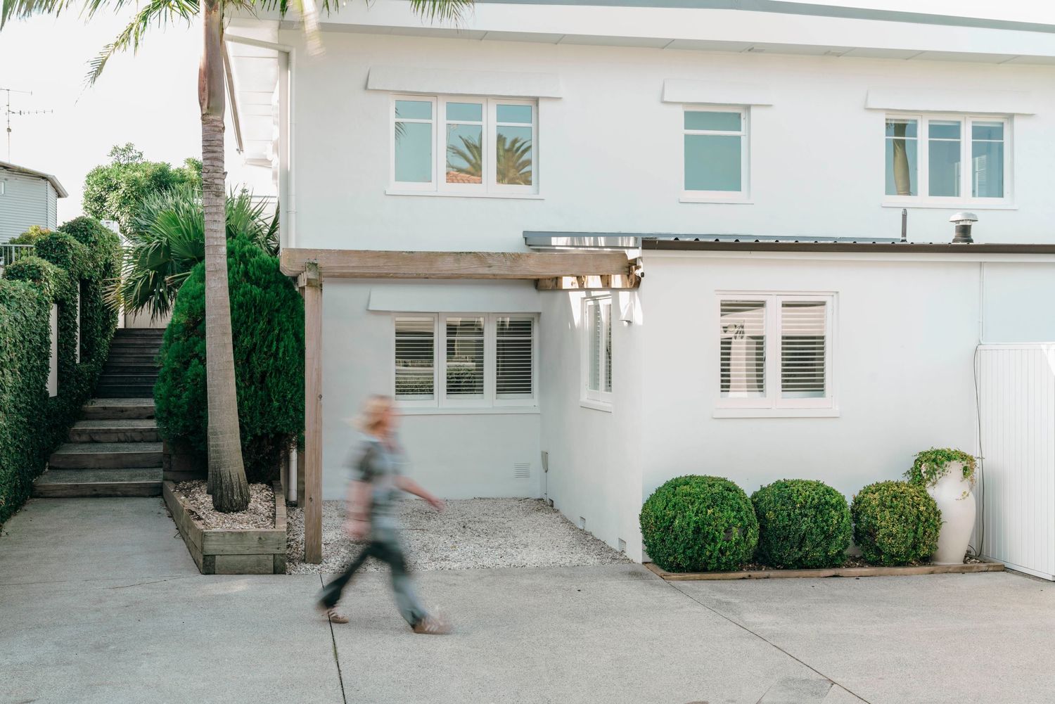 Modern white two-story house exterior with palm trees and round topiary bushes along concrete pathway.