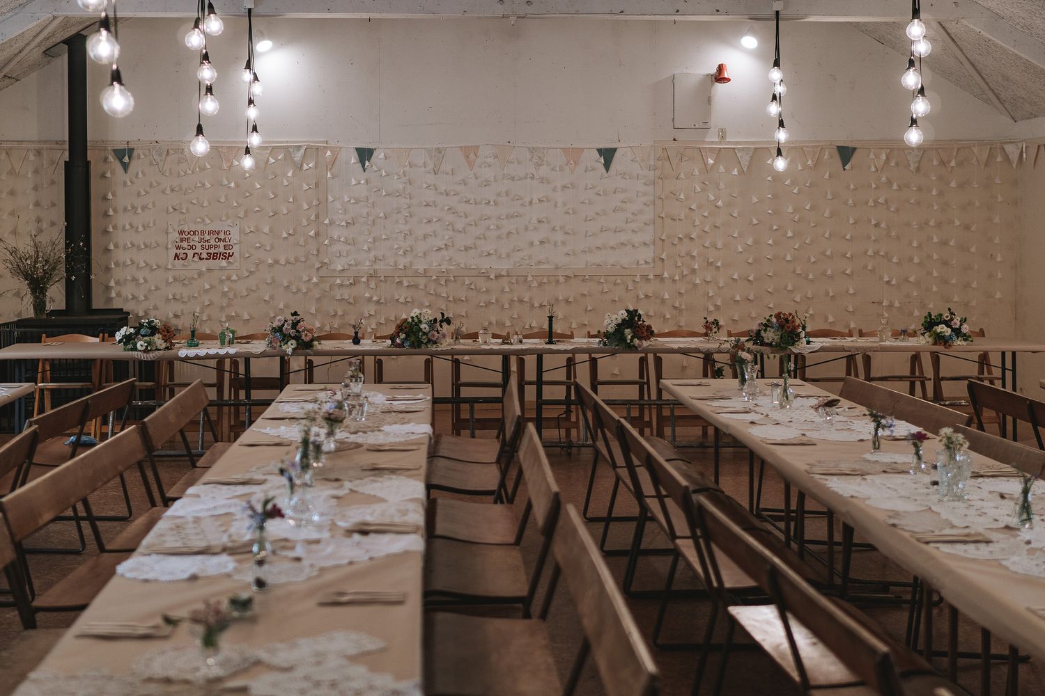 White tables and chairs arranged in rows inside a rustic wedding venue with hanging pendant lights and exposed brick walls.