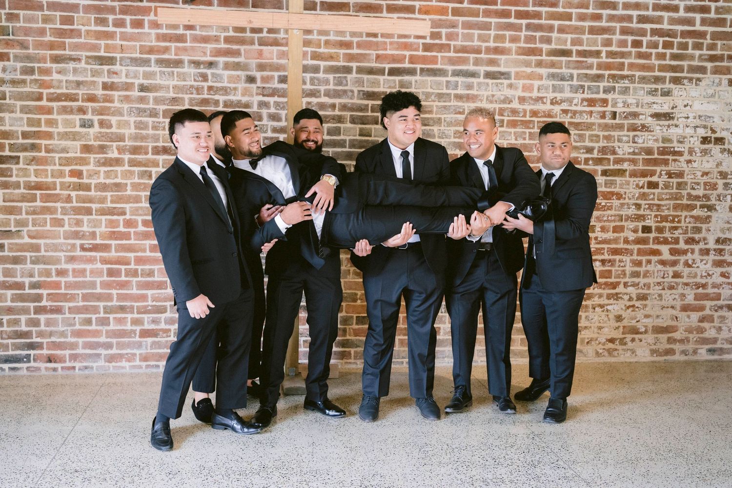 Groomsmen in suits gathered together sharing a laugh against brick wall background.
