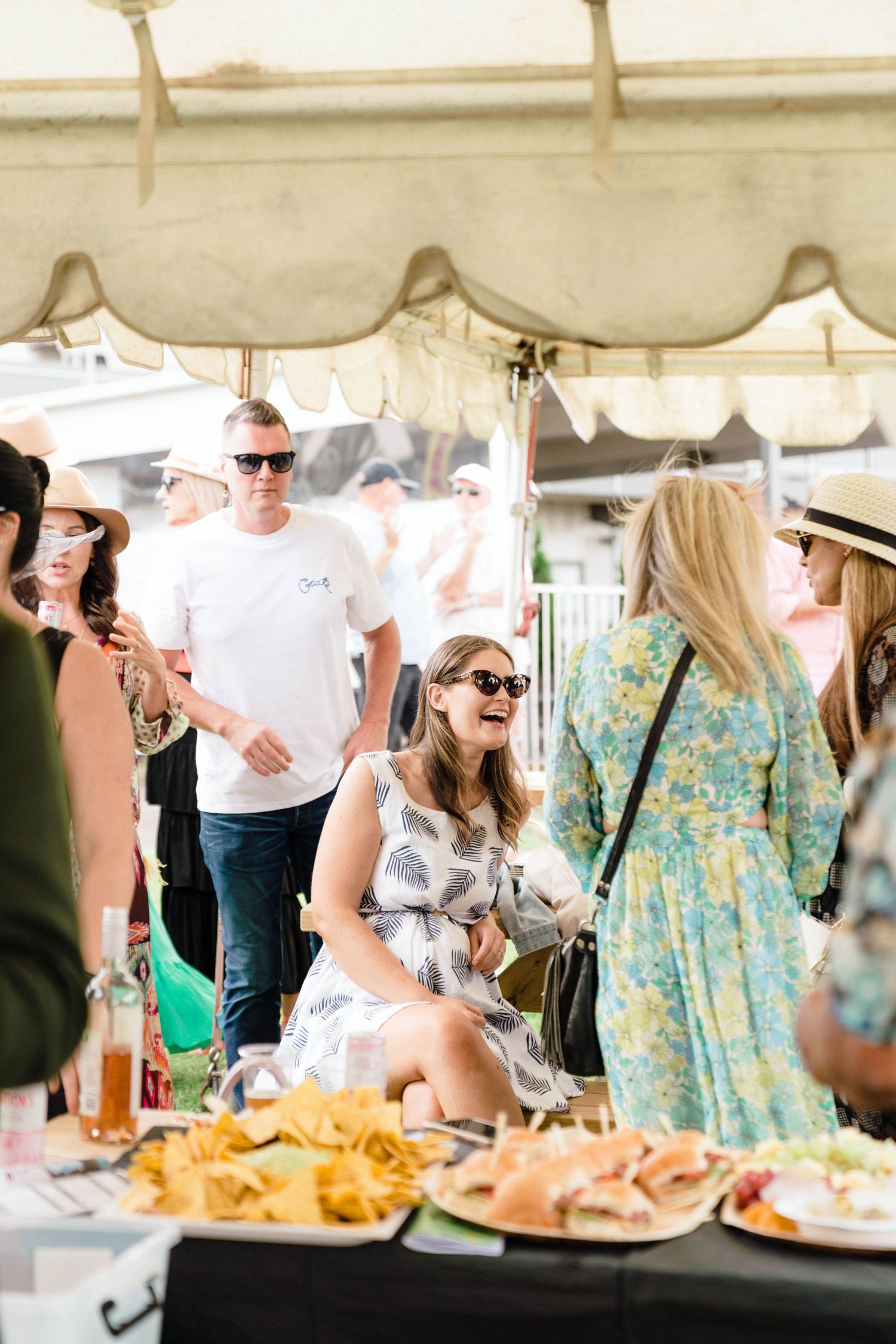 Outdoor party scene with people gathered under a tent enjoying food and drinks.