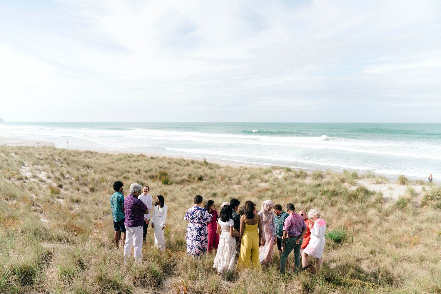 Group of wedding guests standing together on coastal dunes with ocean in background.