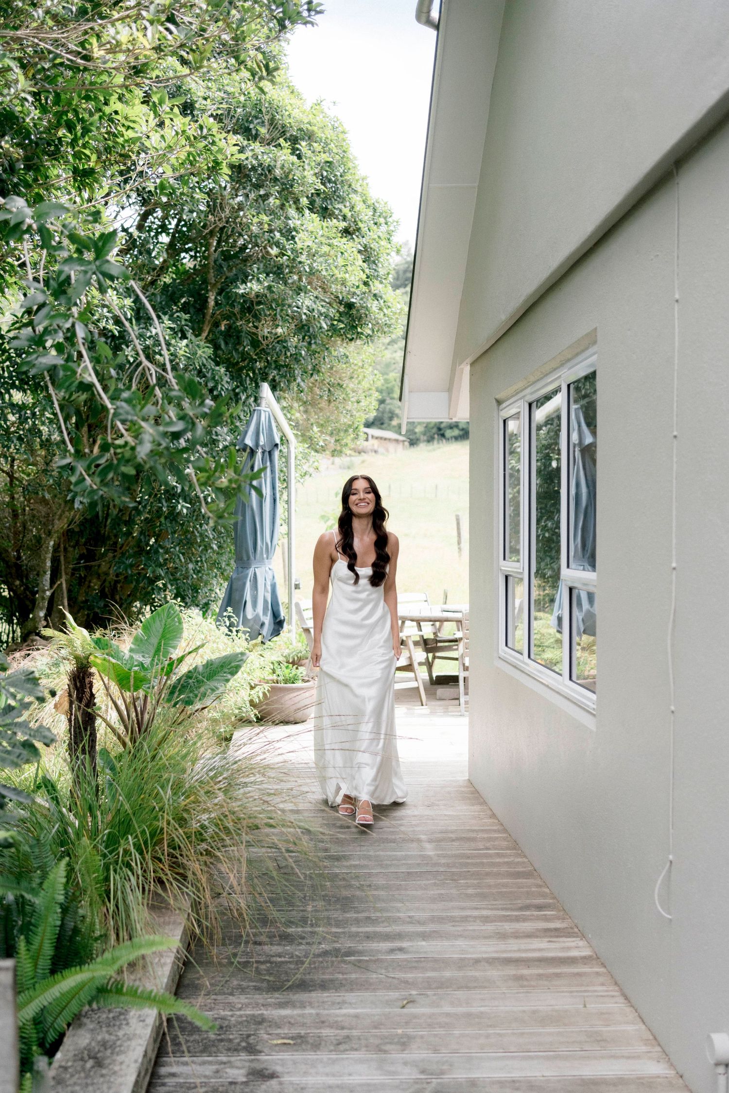 A walkway alongside a white house with lush tropical landscaping and a wooden deck path.