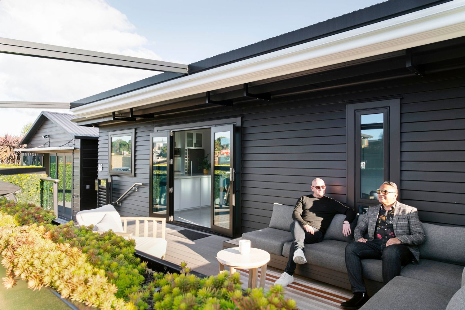 A modern dark gray house exterior with glass doors and a patio featuring people relaxing on outdoor furniture.