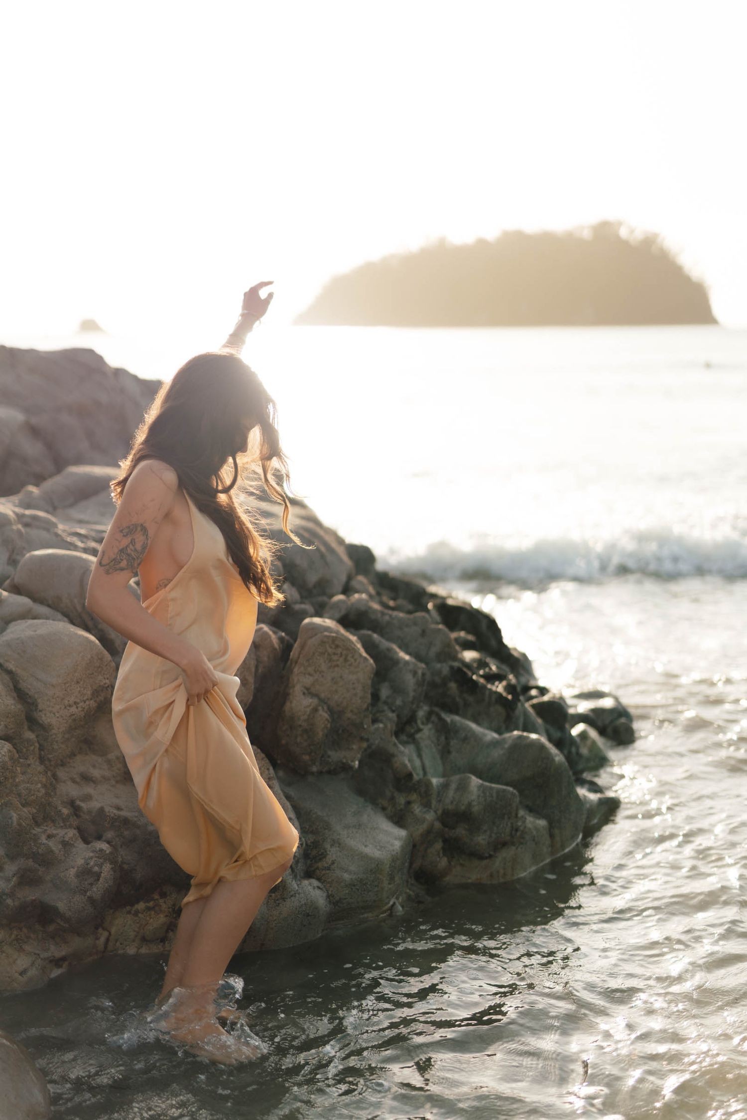 Figure in flowing dress poses dramatically on ocean rocks at sunset.