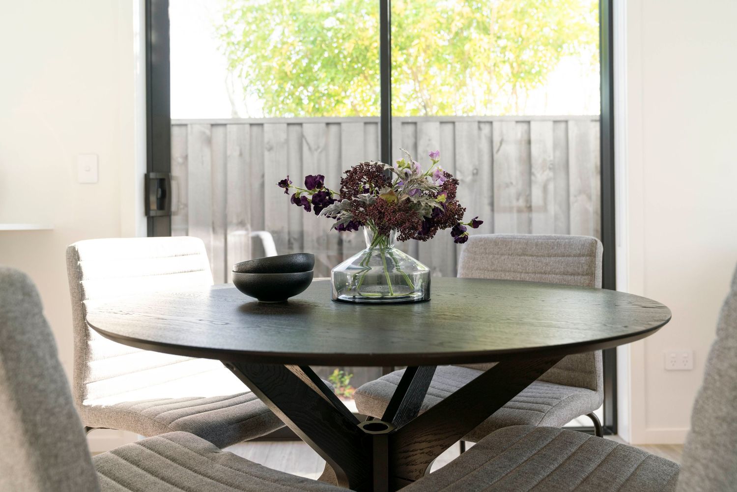 A round wooden dining table decorated with a floral centerpiece and bowl near a bright window.
