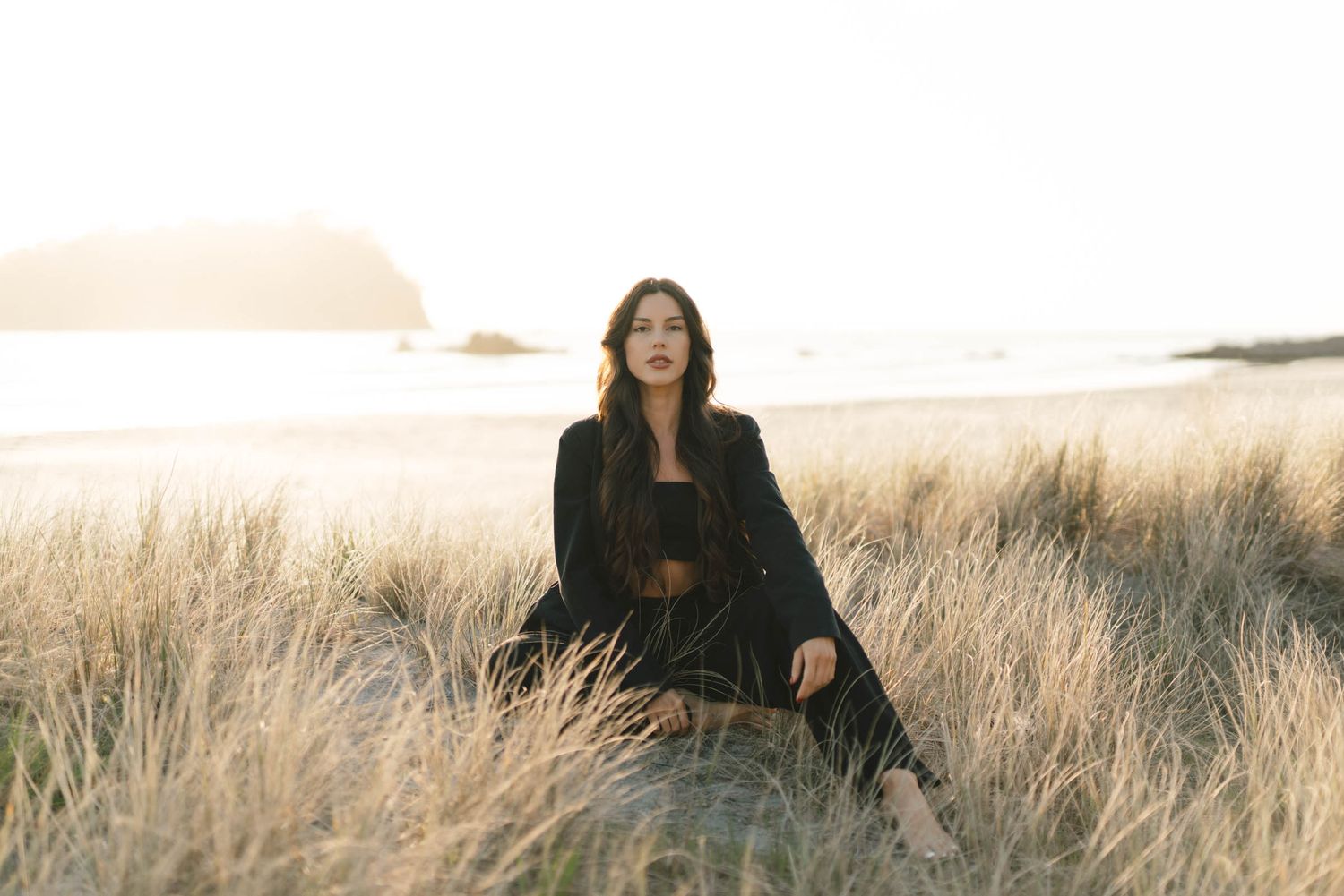 Fashionable black outfit contrasts with golden sunset in coastal grass field.
