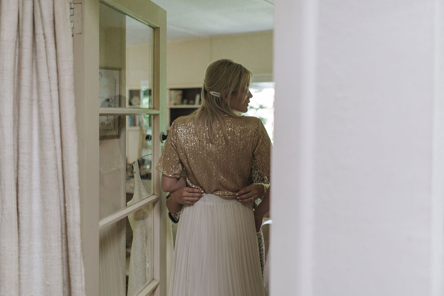 Person wearing a brown sweater and long pleated skirt stands in front of built-in shelving unit in a bright room.