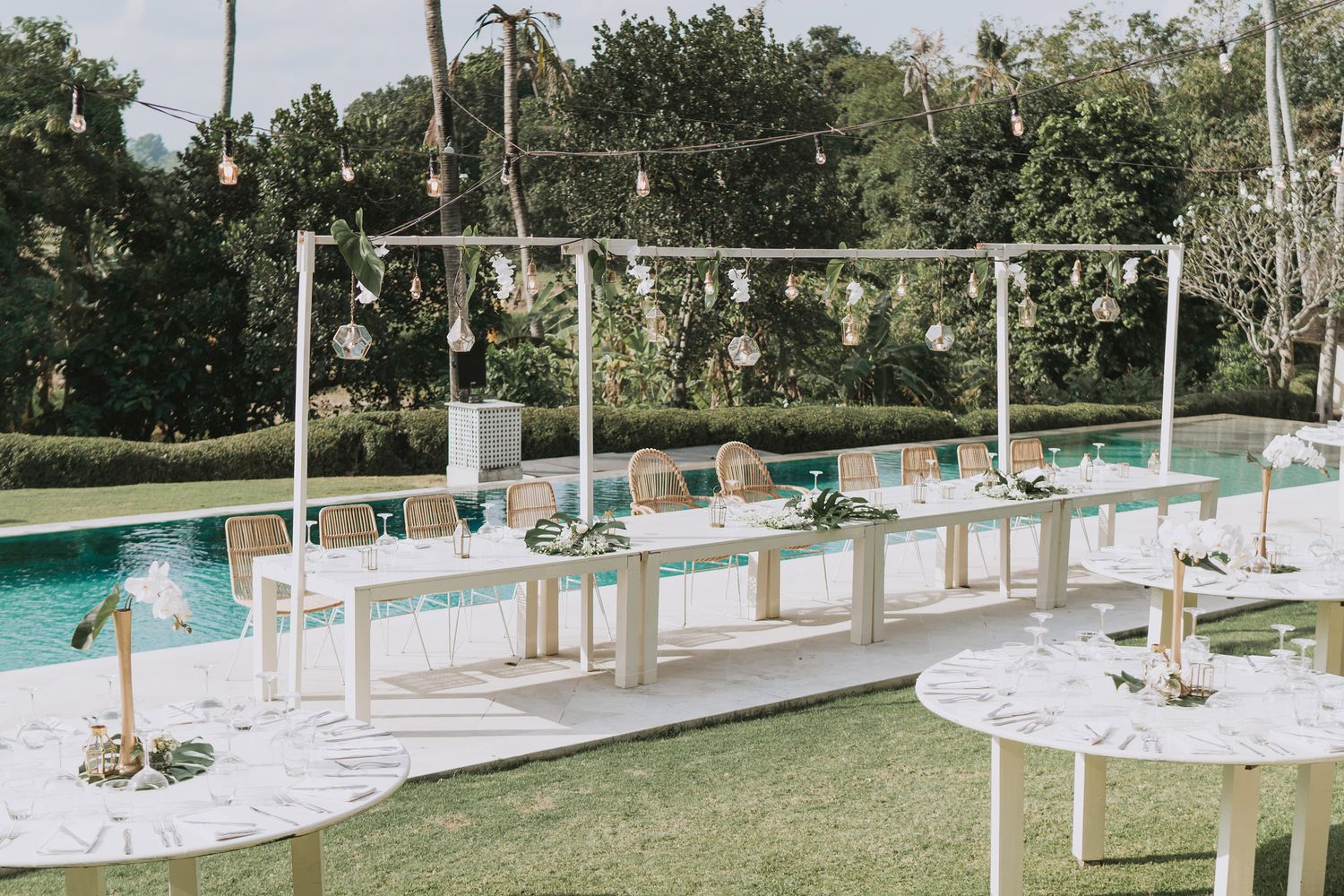 Outdoor wedding venue with white chairs and hanging lights set up along a pool area.