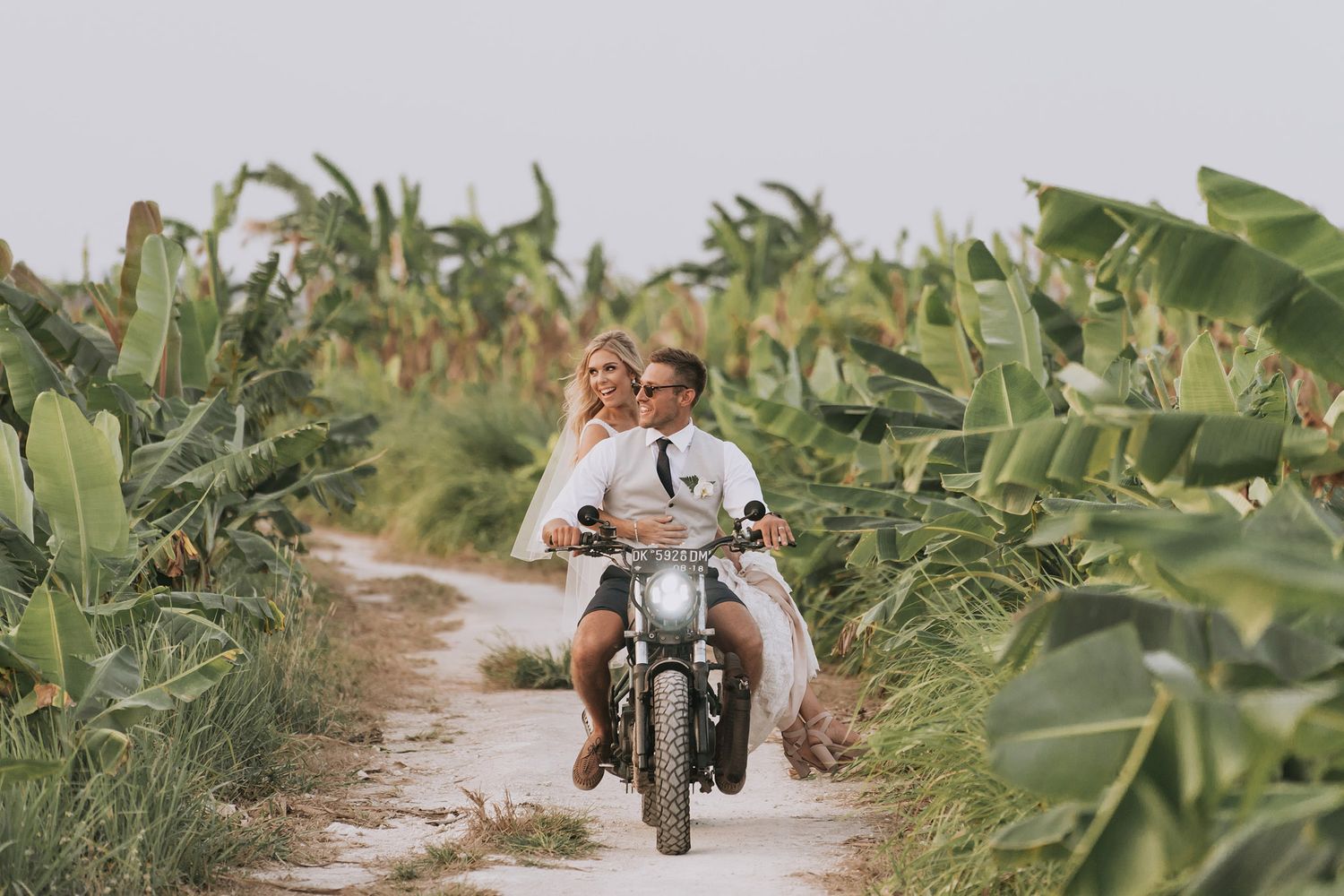 A vintage motorcycle carrying newlyweds rides through lush green foliage on a dirt path.
