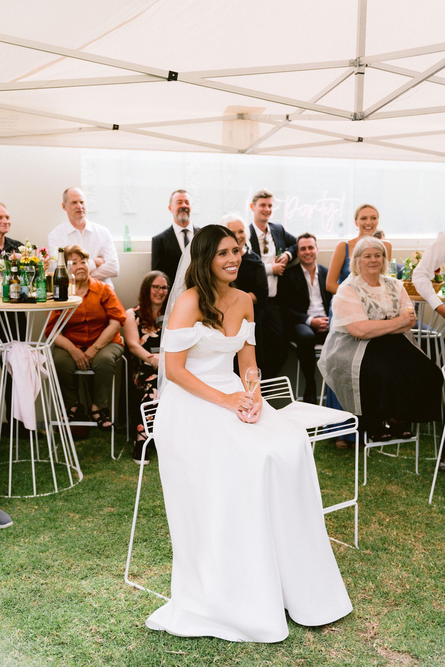 A wedding reception under a white tent with guests seated at cocktail tables.