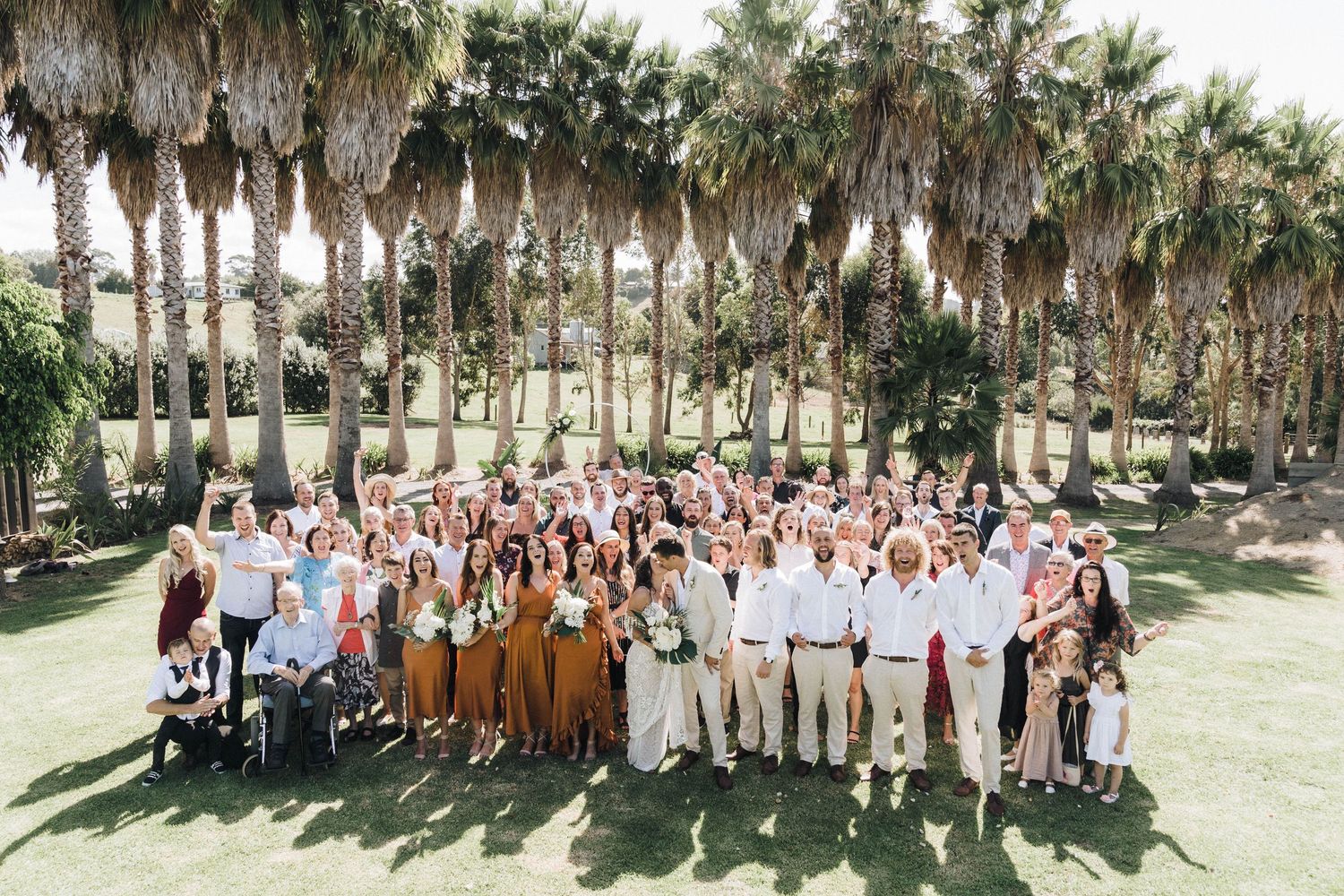 Large group wedding photo with palm trees creating a natural backdrop in a sunny outdoor setting.
